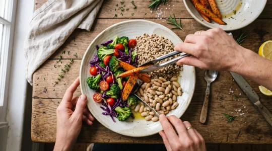 Vue de dessus d'une assiette en cours de composition avec des légumes de plusieurs couleurs, des féculents et des légumineuses répartis par zones sur une table de cuisine en bois.